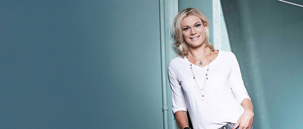 Portr&auml;t einer l&auml;chelnden Frau, Maria H&ouml;fl-Riesch mit blonden Haaren, die an einer Wand lehnt.