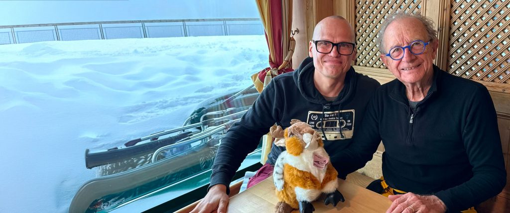 Zwei M&auml;nner, Carsten Neder und Dr. Dirk Hagena sitzen in einer Gondel an einem Tisch, vor ihnen ein Pl&uuml;schtier, im Hintergrund eine verschneite Berglandschaft.