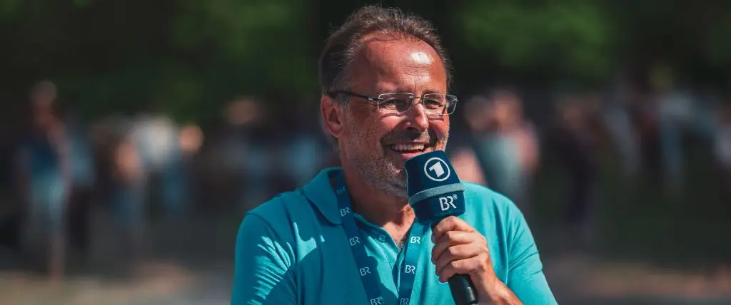 Gerhard &bdquo;Willi&ldquo; Willmann l&auml;chelt mit einem BR-Mikrofon in der Hand vor einem unscharfen Publikum im Freien.